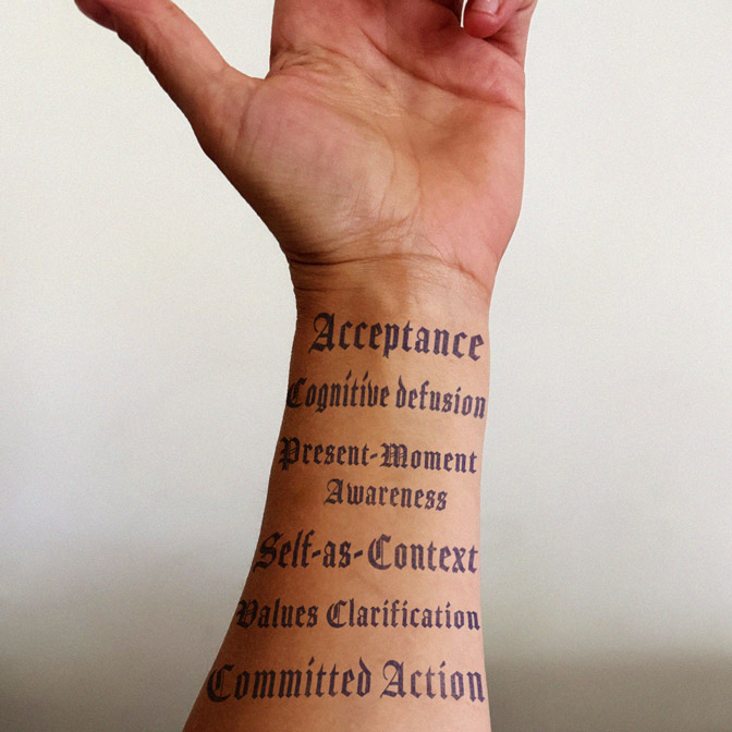 A wrist displaying the six core processes of Acceptance and Commitment Therapy — Acceptance, Cognitive Defusion, Present-Moment Awareness, Self-as-Context, Values Clarification, and Committed Action — in old English script, illustrating ACT therapy concepts.