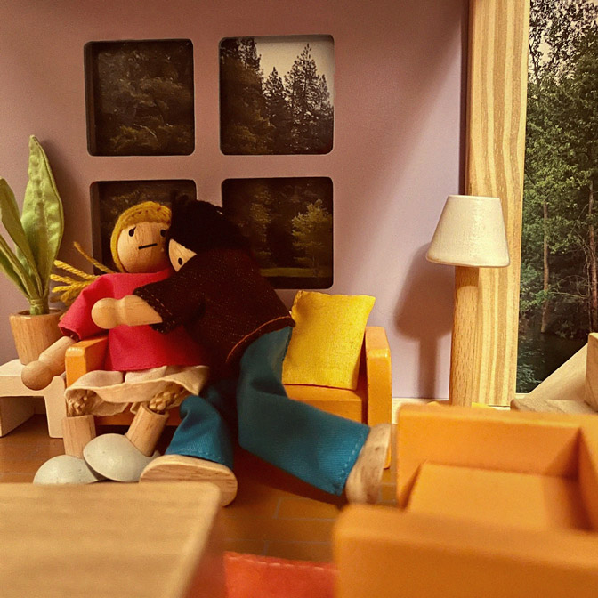 close up of living room set up in a dollhouse with one person leaning over clinging onto the other person on the couch, that doll has a